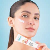Woman applying sunscreen to her face with a tube of sunscreen in front of a blue background
